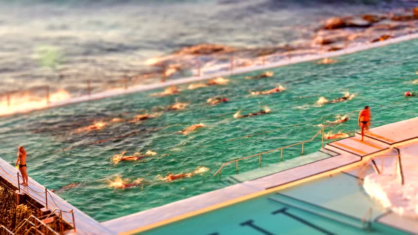 A group of people swims in a large oceanfront pool located right next to the crashing waves of the seathe scene is shot with a tiltshift effect that makes the swimmers and the surrounding architecture look like a miniature model