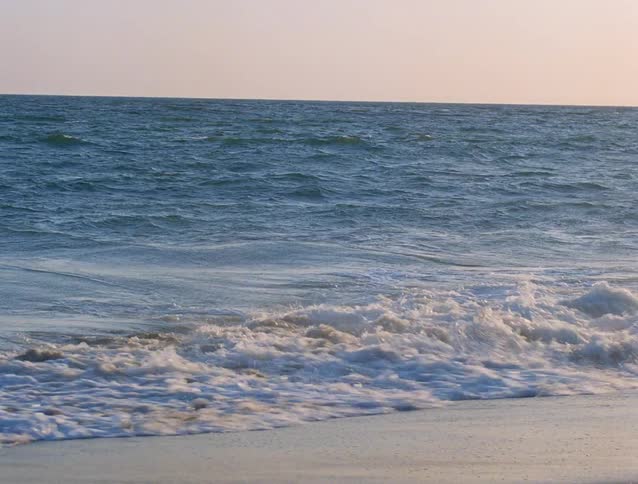 Waves break on the sand under a clear sky on the ocean horizon