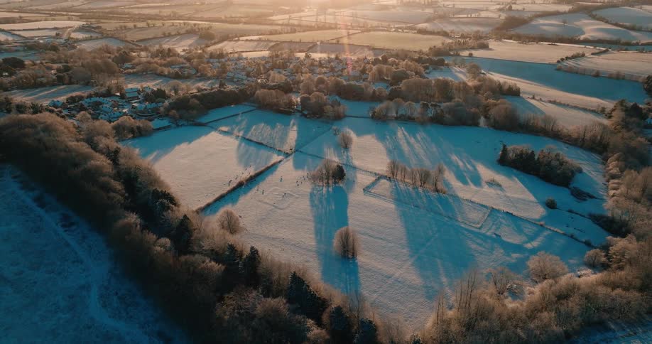 An aerial view shows a vast rural landscape covered in white snow and frost with a small village and long shadows cast by trees during a bright winter morning