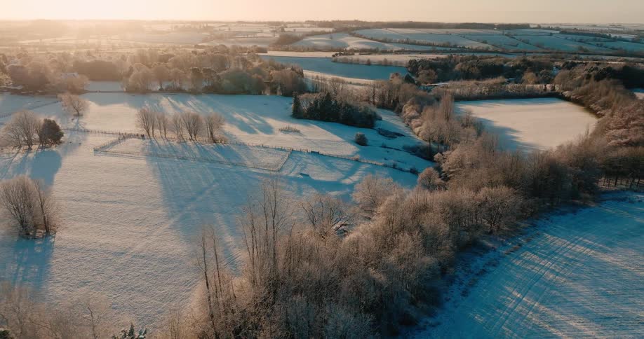 An aerial view reveals frostcovered fields and winding tree lines casting long blue shadows across a serene winter countryside at sunrise