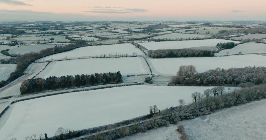 A vast expanse of snow covered agricultural fields and dormant woodlands under a pale morning sky while small country lanes and hedgerows create a dark grid across the frozen white rural terrain
