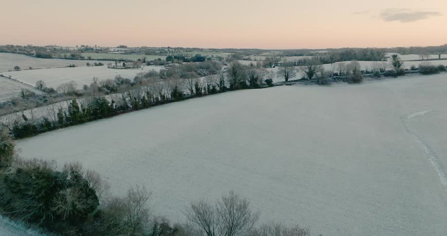 A sweeping aerial view captures rolling countryside blanketed in frost with treelined hedgerows dividing pale winter fields beneath a soft pastel sky