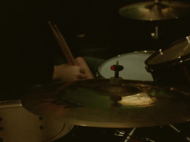 A closeup of a drum kit shows cymbals gleaming under warm stage lighting as a drummers sticks hover midplay in the background