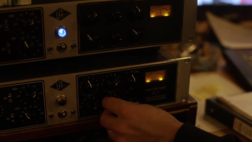 A person turns a black knob on a silver piece of audio equipment with glowing lights in a dimly lit studio