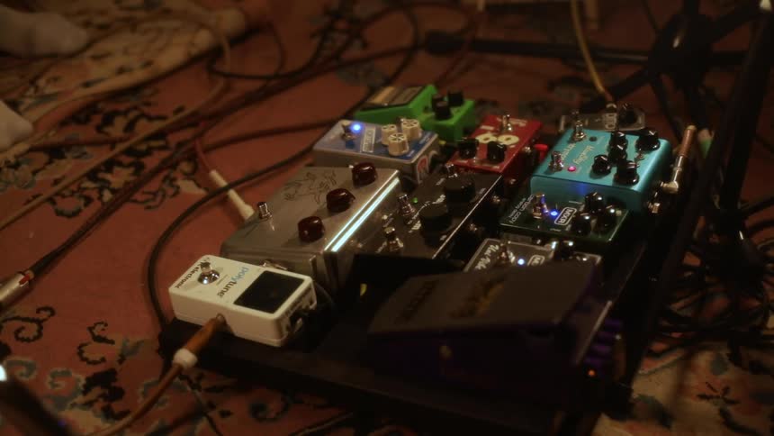 A close view shows a collection of colorful effects pedals for a guitar arranged on a board with glowing lights and messy cables on a patterned rug