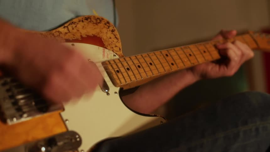 A man wearing a blue shirt and jeans sits down while playing a sunburst electric guitar in an indoor setting