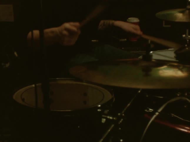 A person with arm tattoos plays a drum set rapidly in a dimly lit setting while a red disposable cup sits on a surface in the background behind the cymbals