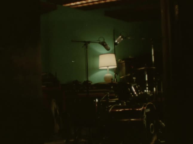 A view through a doorway shows a drum set and a lamp with a glowing shade in a dimly lit music studio with dark green walls