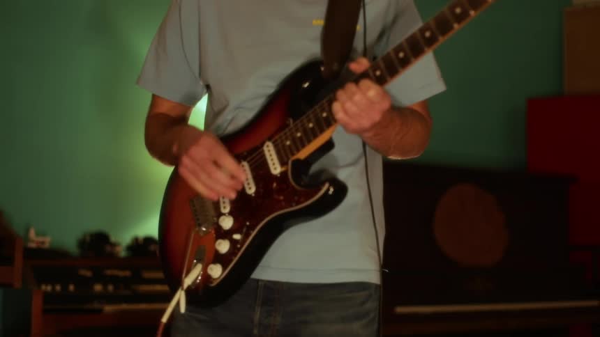 A man in a light blue tshirt and jeans stands in a dimly lit room with green walls strumming a sunburst electric guitar