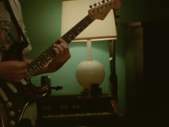 A person from the shoulders down is show standing playing an electric guitar and a flash image shows a man seated playing the drums in a dimly lit room