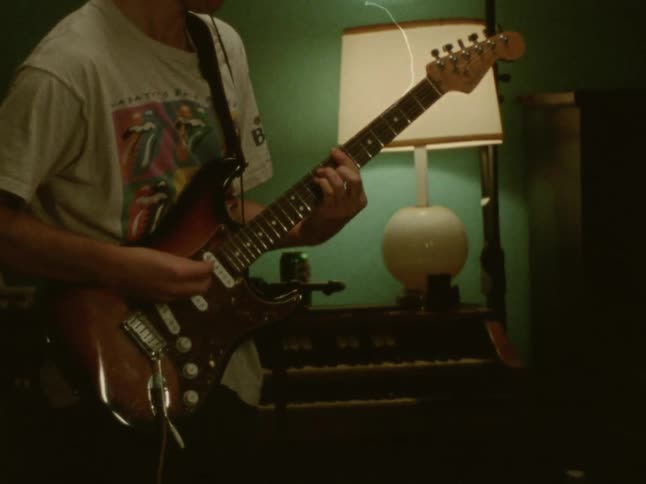 A man wearing a graphic tshirt plays an electric guitar in a room with a lamp and a piano in the background
