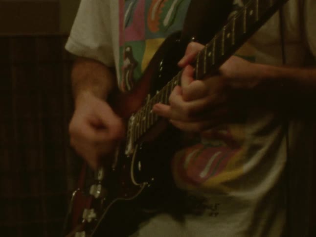 A close view of a person wearing a graphic shirt playing a dark sunburst electric guitar in a room with a lamp