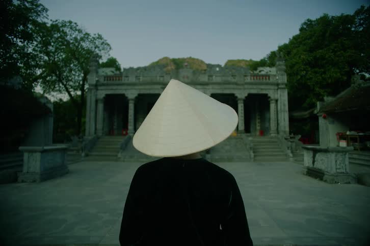 A person wearing a traditional conical hat stands facing an ornate stone temple framed by trees and hills in the background