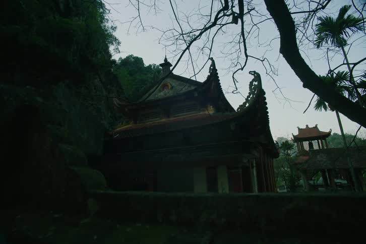 A traditional asian temple with an ornate curved roof sits nestled against a steep rocky cliff under a dim sky