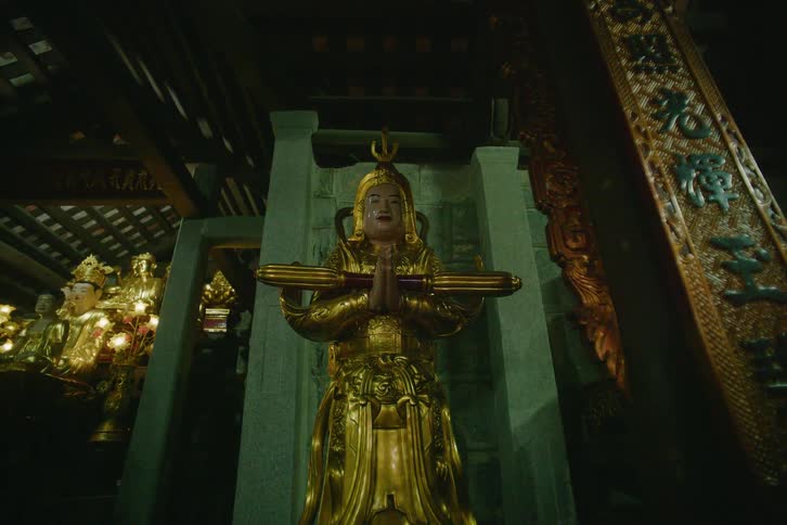 A gilded statue of a serene figure stands within an ornate temple interior hands pressed together in prayer beneath intricate wooden beams and carvings