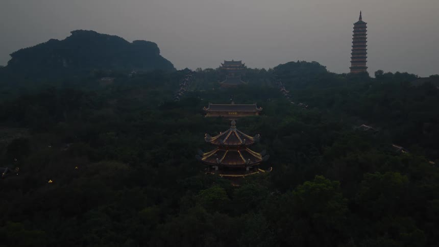 An aerial evening shot showcases a sprawling complex of traditional east asian temples and a tall pagoda nestled within a dense dark forestthe structures are illuminated with golden lights that contrast against the twilight shadows and the silhouettes