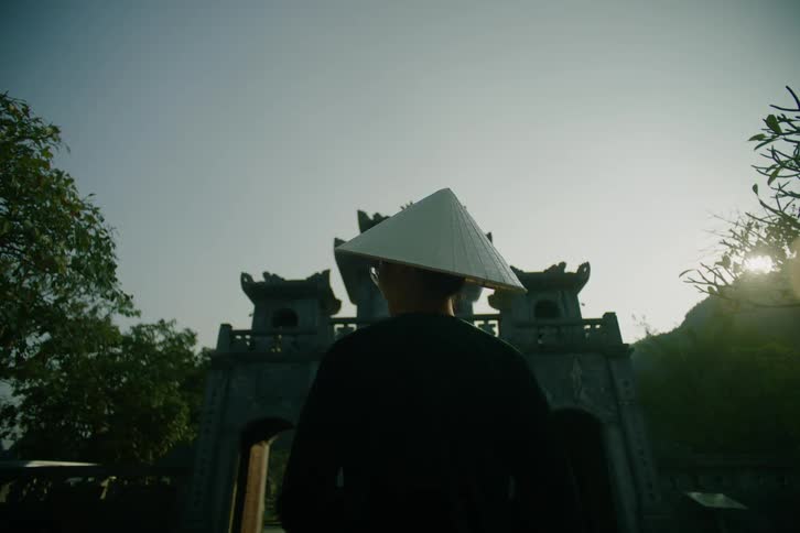 A man wearing a traditional conical hat stands with their back to the camera while looking toward a large stone gate at a temple during the day under a clear sky with trees and mountains visible in the background