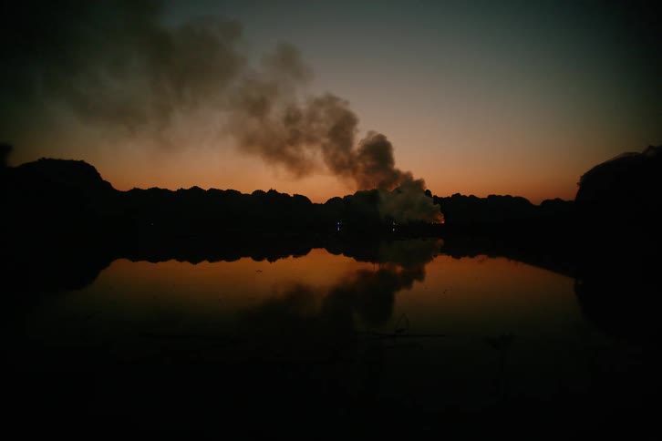 A dark plume of smoke rises from a fire behind a row of hills and reflects on the surface of a calm lake under an orange and blue twilight sky