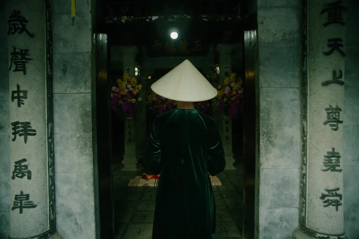 Asian female wearing a rice hat standing at the door of a temple with an altar surrounded by colorful flowers indoors during day time