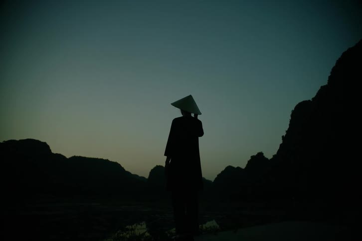 A lone figure in a conical hat stands silhouetted against a fading twilight sky between dark mountain ridges
