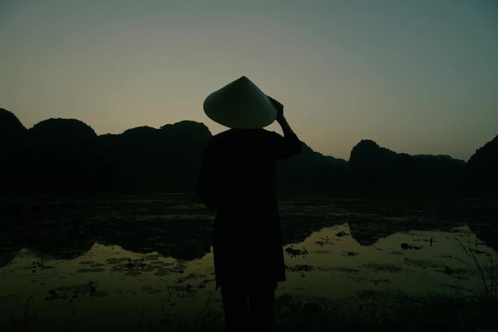 A silhouetted figure wearing a conical hat stands by a reflective body of water against a backdrop of dark mountains at sunset