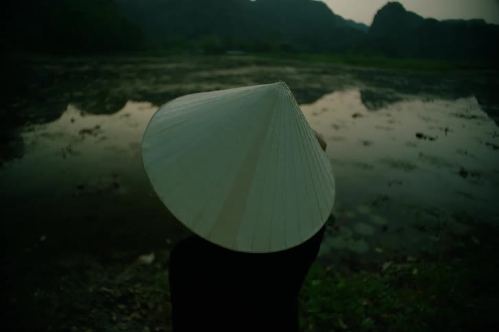 Female asian person wearing a rice hat standing in front of a pond with hills on the background outdoors during dusk or dawn