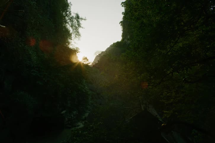 Bright golden sunlight shines through a narrow opening between tall dark cliffs covered in green trees with a warm flare effect across the scene