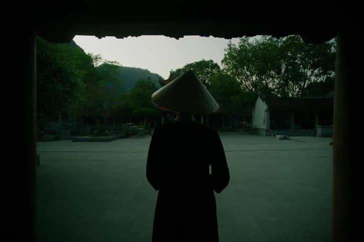A woman wearing a traditional conical hat stands in silhouette within a dark doorway looking out toward a stone courtyard and lush green mountains under a dim evening sky in a quiet rural setting
