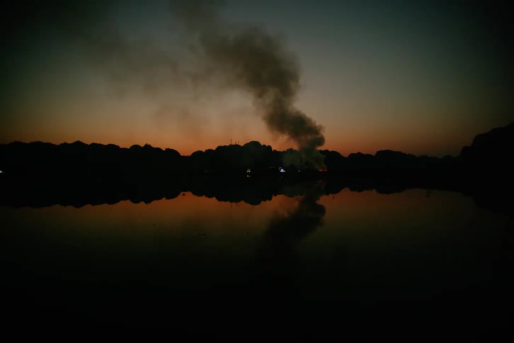 Smoke rises into a colorful dusk sky from a small fire beside a calm lake its reflection mirrored in the still water