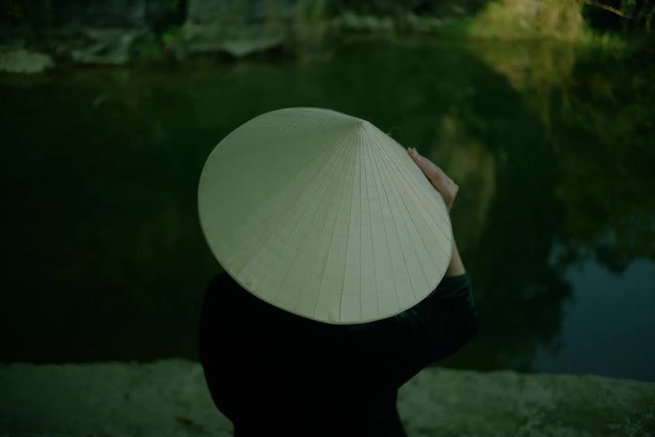 A woman wearing a traditional conical hat sits with her back to the camera while looking out over a calm dark river surrounded by rocky banks and green trees in a quiet natural setting