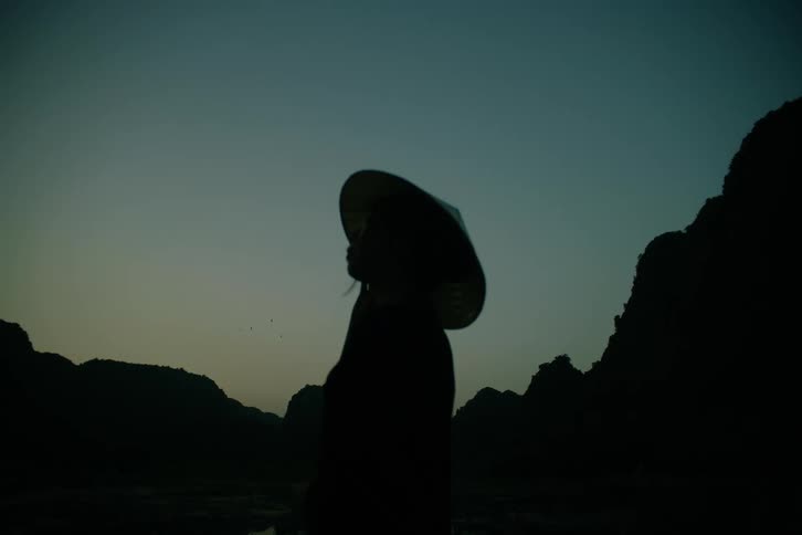 A person wearing a traditional conical hat stands in silhouette against a darkening twilight skybehind them the jagged outlines of a mountainous landscape fade into the deep blue of the evening