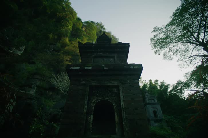 An ancient stone temple doorway stands nestled against a lush forested hillside silhouetted against the pale morning sky