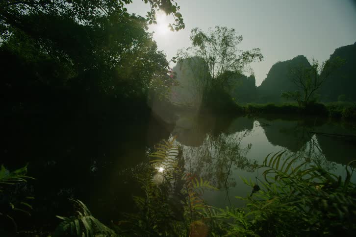 The bright morning sun hangs over a calm river reflecting tall limestone mountains and lush green jungle foliage while ferns and bamboo frame the serene tropical landscape in a wide shot