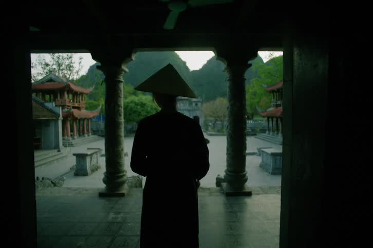 Silhouette of an asian female wearing a rice hat standing at the porch of a historic building with a forest and hills on the background