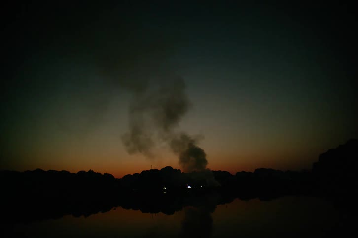 Water reflects nearby trees and smoke rising from a fire at night