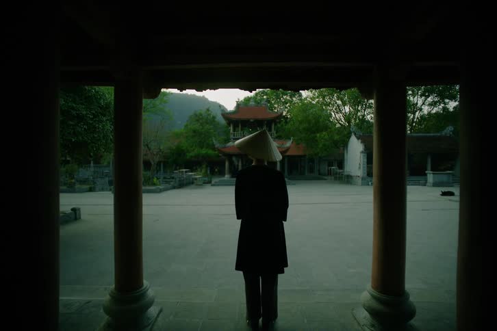 A person wearing a dark coat and a traditional conical hat stands still in a courtyard framed by dark pillars looking toward a stone temple and green trees