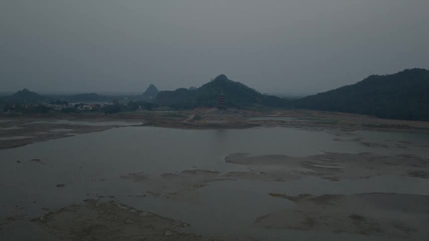 A distant aerial view shows a red pagoda standing on a hill surrounded by a vast empty lake bed and dark mountains under a hazy grey sky