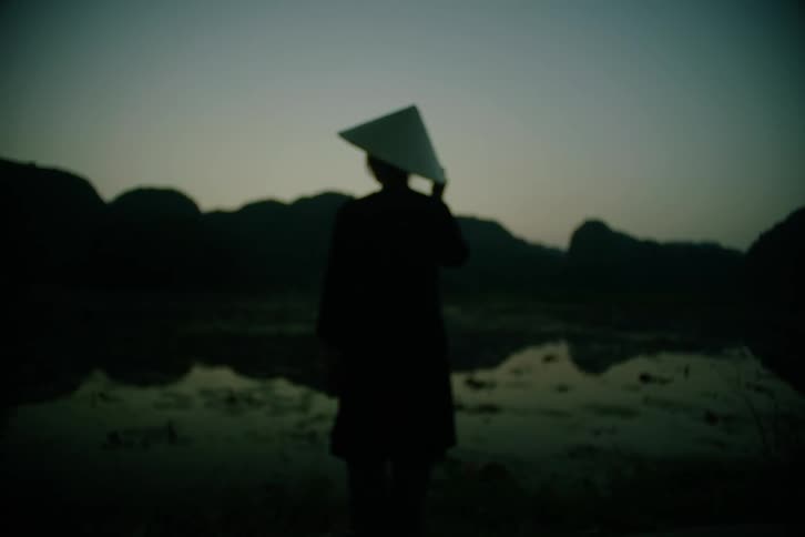 Silhouette of an asian female wearing a sun hat standing in front of a pond outdoors during dusk or dawn