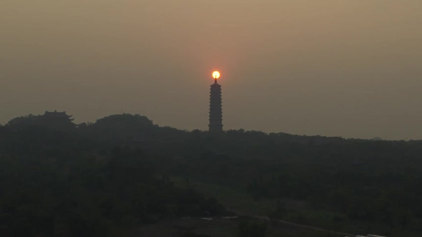 The sun aligns perfectly atop a tall pagoda rising above a darkened tree line at dusk