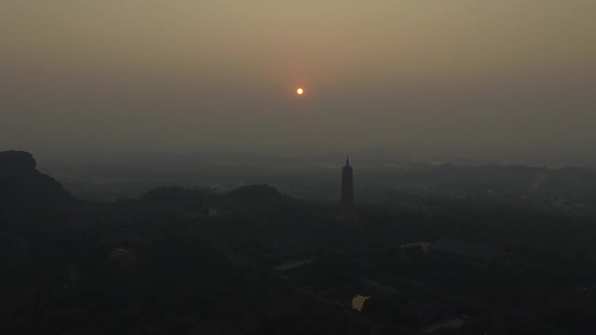 A tall pagoda stands silhouetted against a hazy sky as the orange sun sets over a vast forest and distant mountains