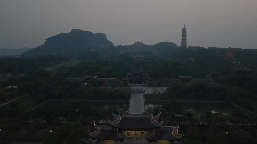 An aerial view reveals an expansive temple complex nestled among lush greenery and limestone mountains at duska tall pagoda and traditional curved roofs stand out as the camera glides toward a central gate and bridge