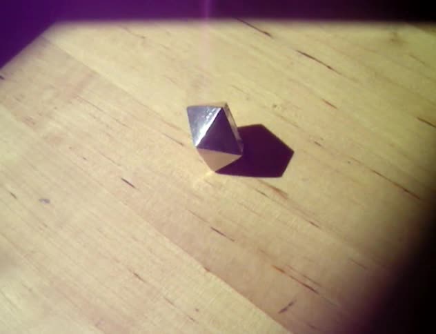 An angular geometric and metallic object is spinning on a wooden floor with sun flares