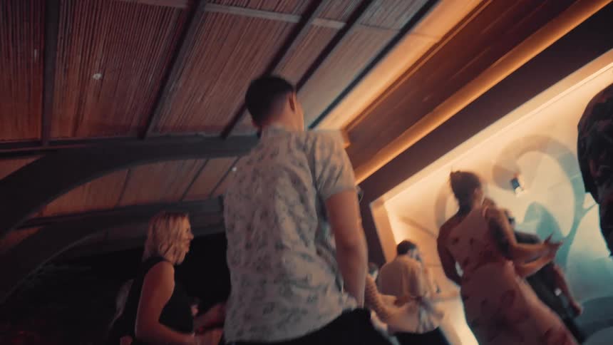 A group of people are seen dancing together in a dimly lit indoor space with a rustic ceiling the camera focuses on a man in a patterned shirt moving to the music amidst the crowd