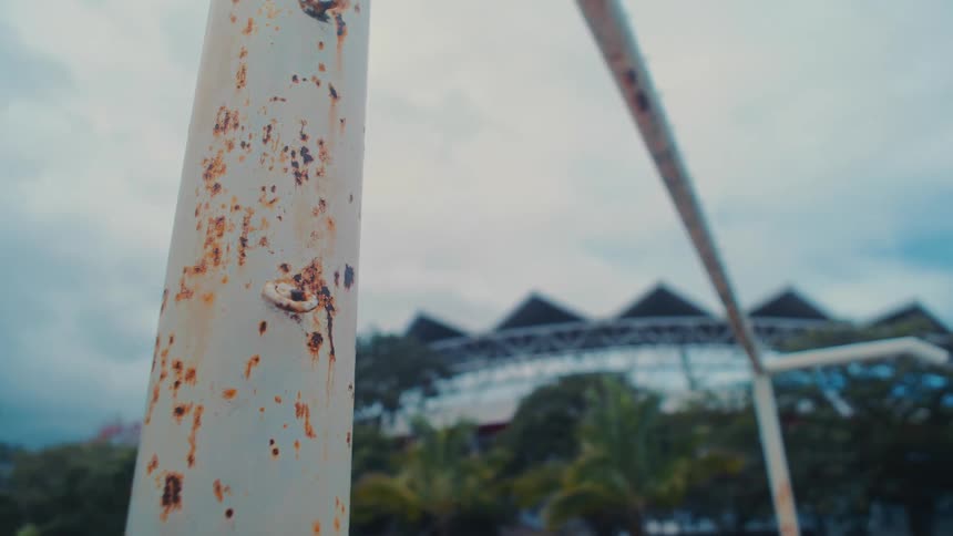 A football goalpost made of steel with rust on its surface stands in an open field near a stadium