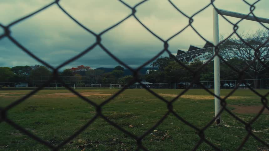 The video features a view of a grassy soccer field and goalpost seen through a chainlink fence under a cloudy sky the camera pans slowly to the right highlighting the empty sports complex and surrounding trees in a moody lowlight setting