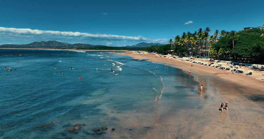 People walk on the beach and in the water as palm trees overlook the coast with mountains in the distance