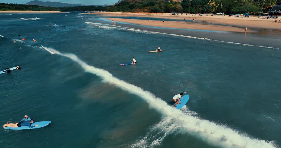 Surfers dot the blue ocean waters near a sandy coastline with several individuals paddling or sitting on their boards while waiting for waves one surfer successfully catches a wave and rides it toward the shore as the white foam trails behind them
