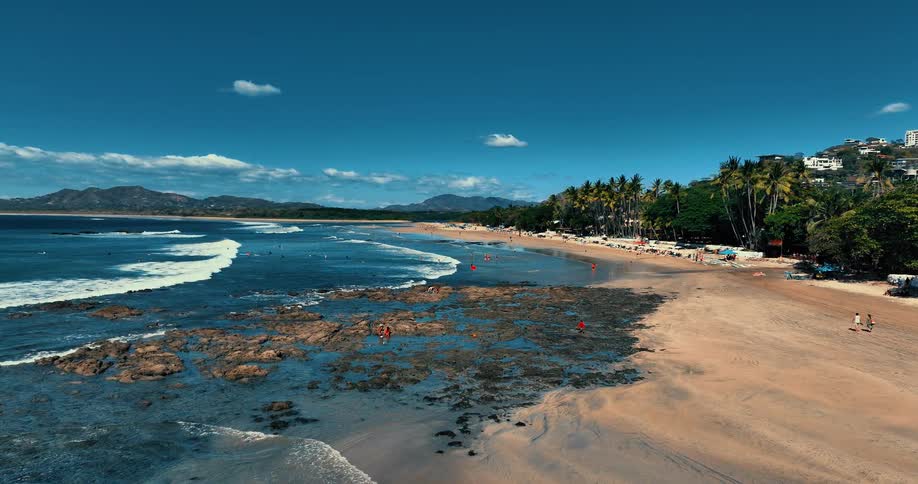 The video shows an aerial drone shot of a tropical beach where gentle waves roll onto a sandy shore lined with palm trees and scattered crowds in the foreground rocky tide pools are exposed at low tide with people exploring the waters edge under a cle