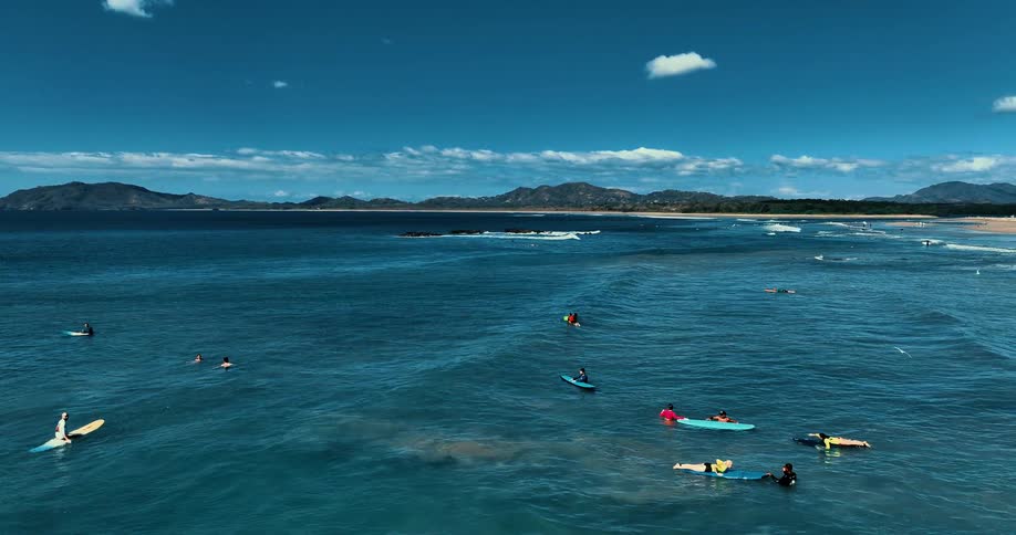 Aerial view of surfers and swimmers in the ocean near a beach with mountains in the background