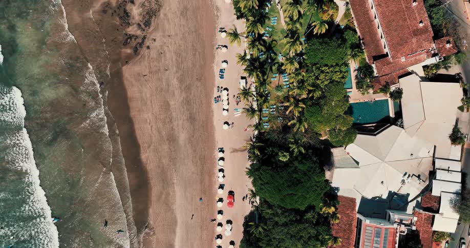 Aerial view of a neighborhood along the coast with people walking by umbrellas on the beach as waves roll in to shore and cars passing buildings on the street